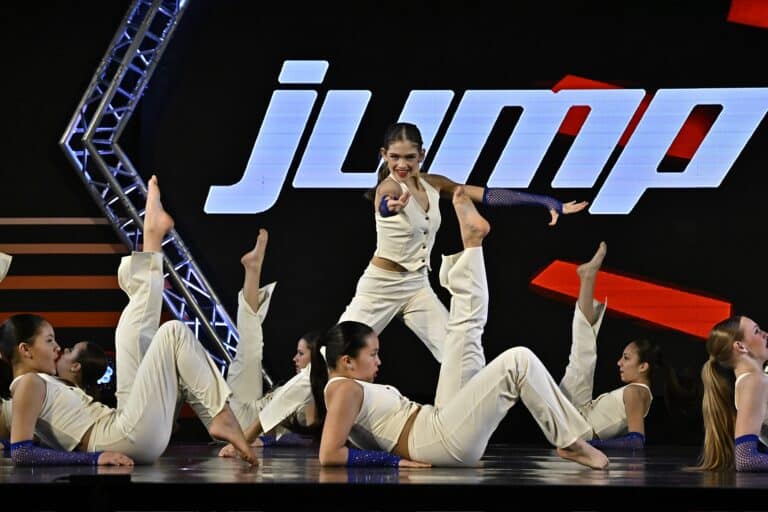 Dancers performing a choreographed routine in white costumes on stage with a bold "jump" backdrop.