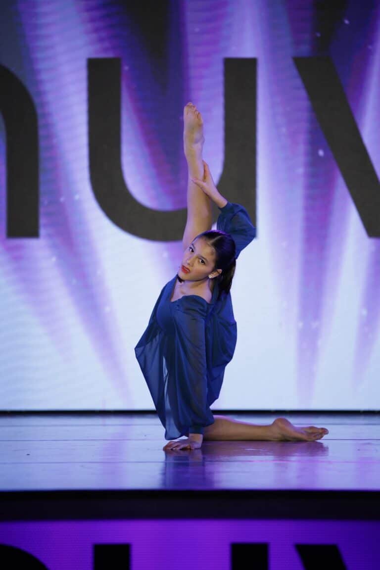 Solo dancer in a blue costume performing a floor pose with a vertical leg extension on stage at the Nuvo Dance Convention 2025.